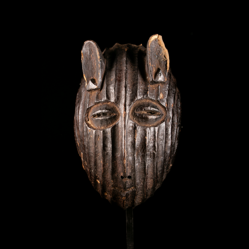 Front view of a Metoko wooden african mask with vertical carved grooves displayed on its custom metal stand.